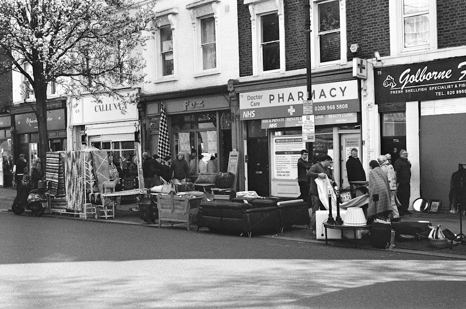 A black and white photo of a storefront featuring a large, intricately patterned Persian-style rug hanging in the window, with a collection of smaller, patterned rugs draped on the right side. The rug