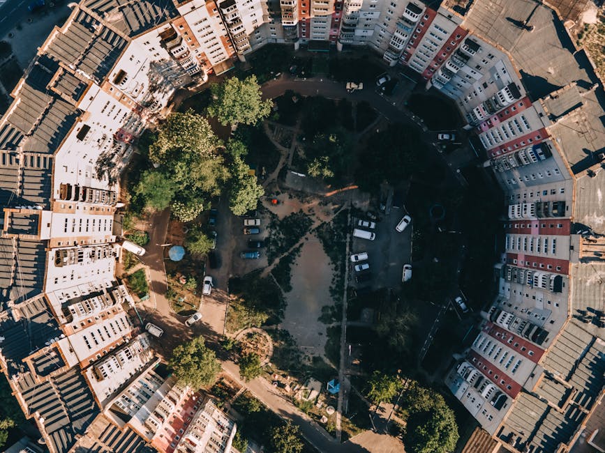 An aerial view of a residential courtyard surrounded by multi-story apartment buildings with various colored facades including white, beige, pink, and gray. The courtyard features paved walkways, gree